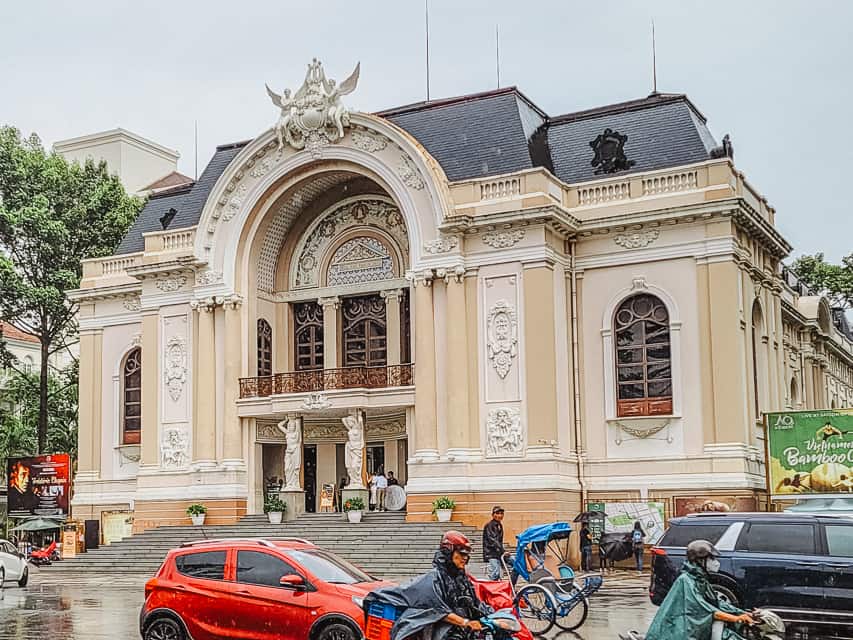 A cream opera house with detailed etchings and statues on the side of a busy street with motorcyles and cars.