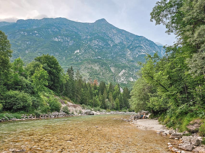 A clear water, shallow, river spreads out to both sides of the beach with trees and mountains on both sides.