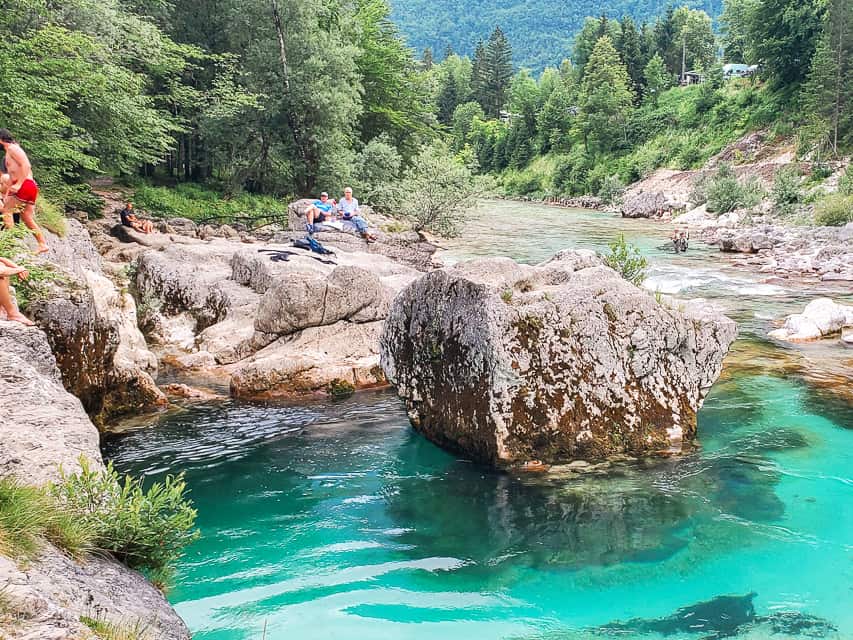 People sit on the beach of the soca River with rocks on both sides and crystal clear blue water everywhere