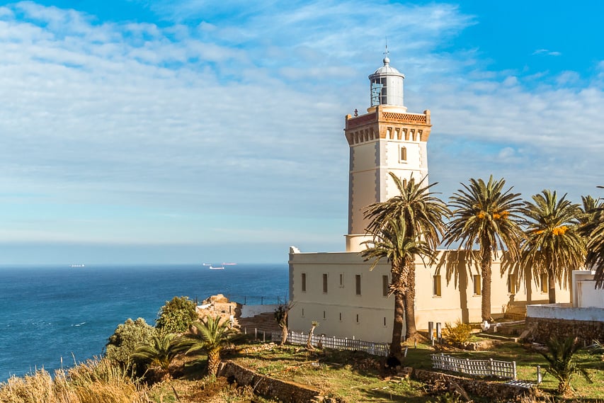 A white stone lighthouse stands on the edge of the moroccan coast with beautiful blue waters and skies.