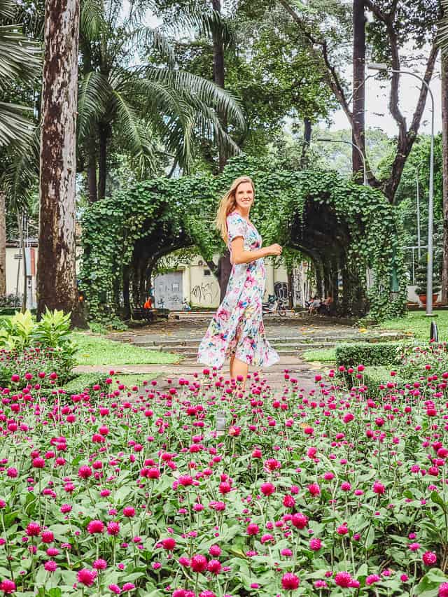 A woman in a floral dress stands in front of pink carnations and a fern gateway.