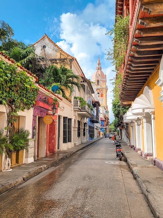 A colorful alley way with lots of different colored doors and plants. There is an intricate steeple in the background