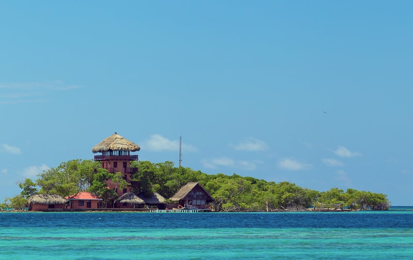 A luxurious island with a large wood house with a large balcony looking out at the deep blue / teal water.
