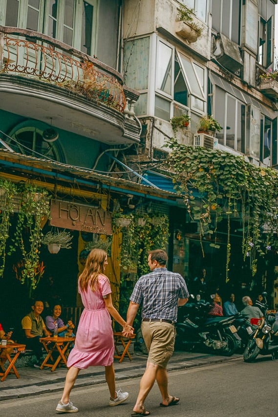 A man and woman walk in the streets of a cute shop filled alley with vines coming over the balconys.