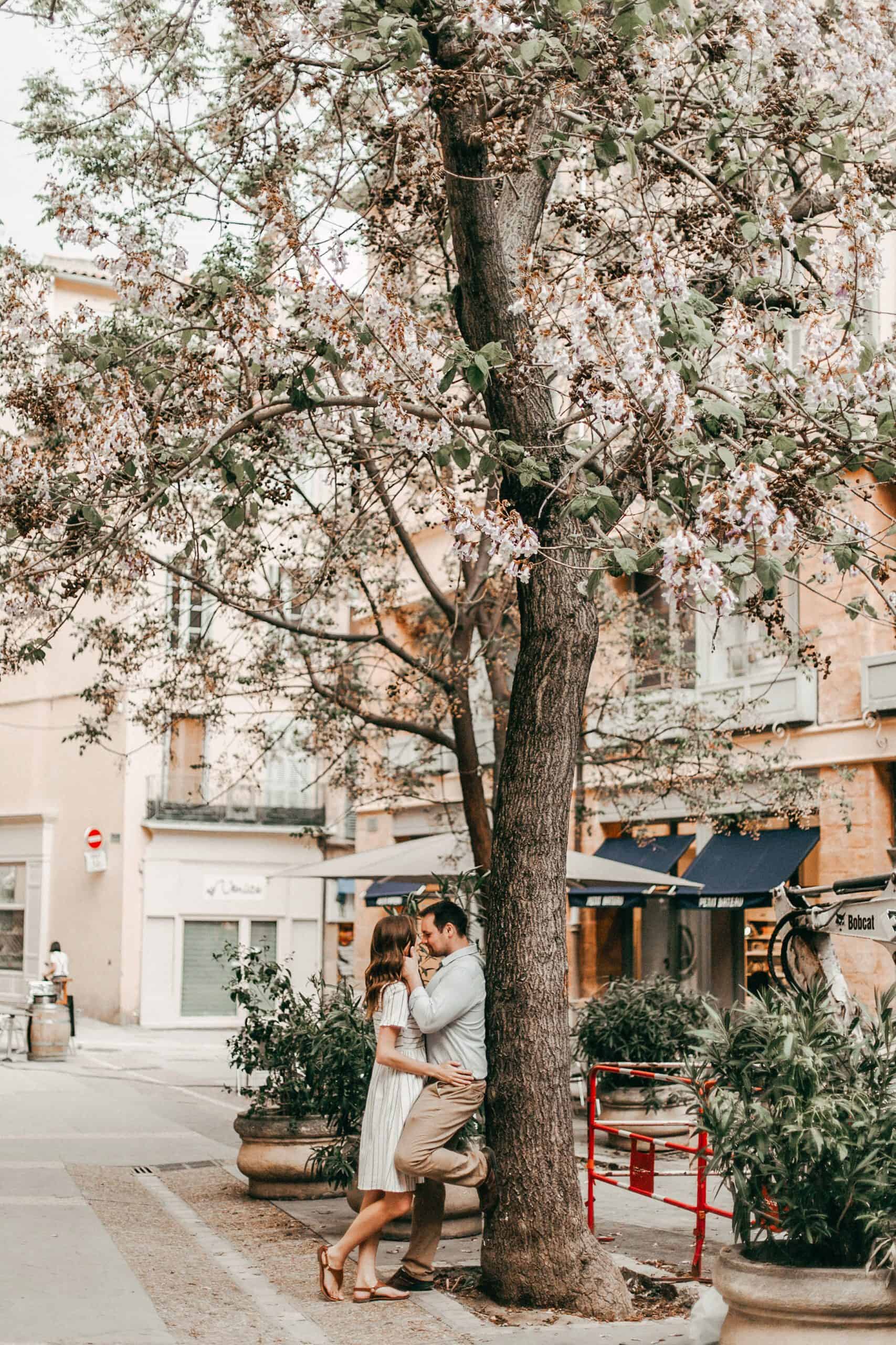 A man and woman kiss leaning next to a flowering tree on the side of a sidewalk with lots of plotted plants/bushes on the left and right of them.