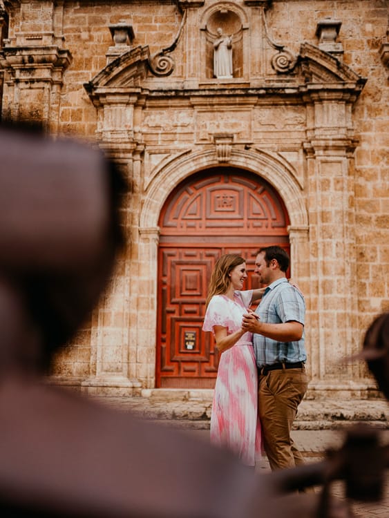 A man and woman dance on the front of a brick and stone church with rusting metal artifacts on the other side of the street.