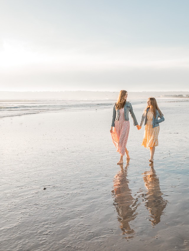 A daughter and mom walk on the shoreline of a blue water beach with the sun starting in to set in the background.