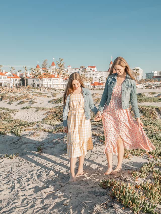 A mom and daughter hold hand walking on a sandy beach with gorgeous white and coral houses behind them