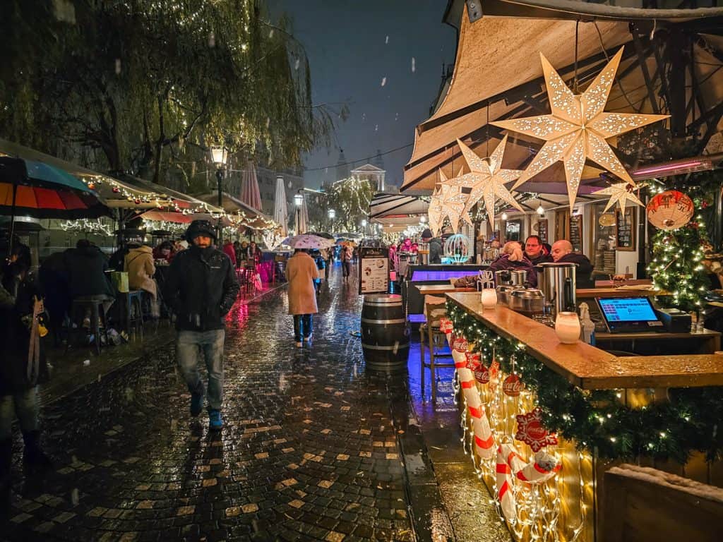 A sidewalk with lots of string lights next to small huts with the Europe paper stars with intricate designs that are lit up.