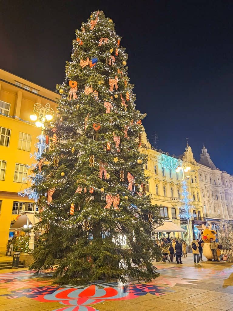 A large Christmas tree almost as tall as the buildings in the Zagreb Christmas market is decorated with large bows and gold ornaments.