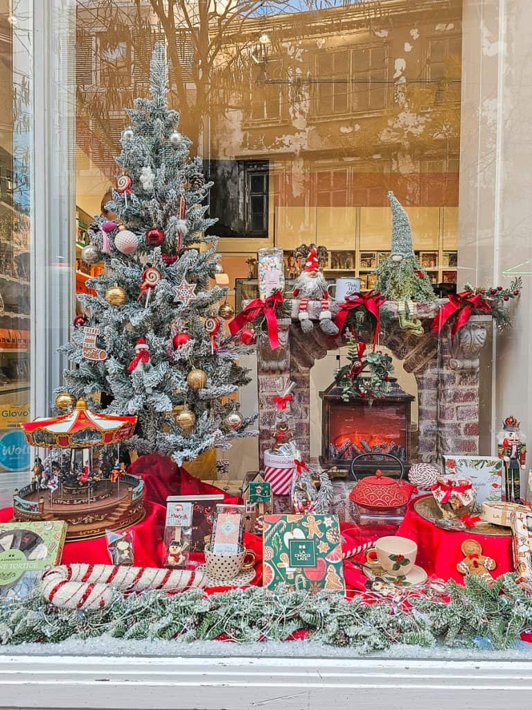 A large assortment of Christmas decorations like cookbooks and teacups sit on a stand next to a fireplace and Christmas tree