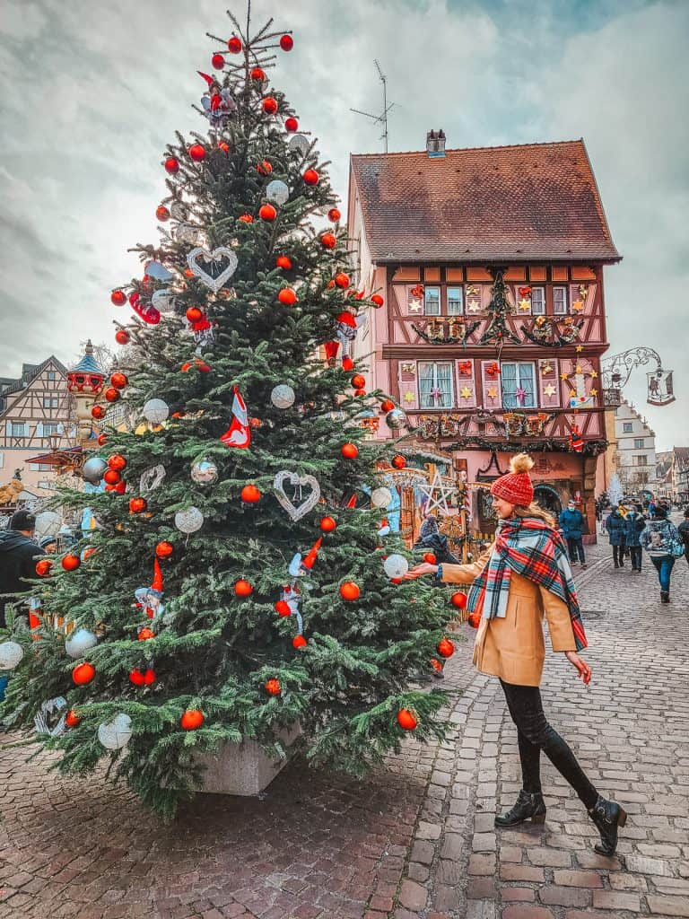 A woman holds one of the white ornaments on the Christmas trees. There are red ornaments on the tree matching the bows on the pink houses behind the tree.