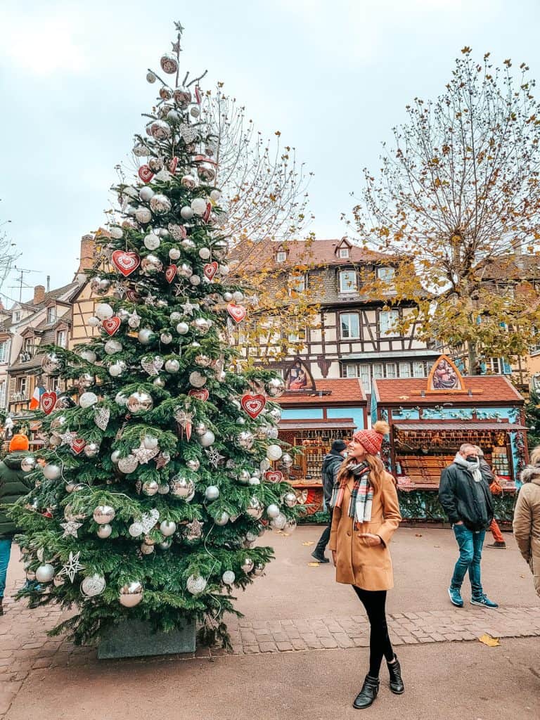 A woman stands to the side of a wooden Christmas market stall with blue trim and little mugs being displayed.