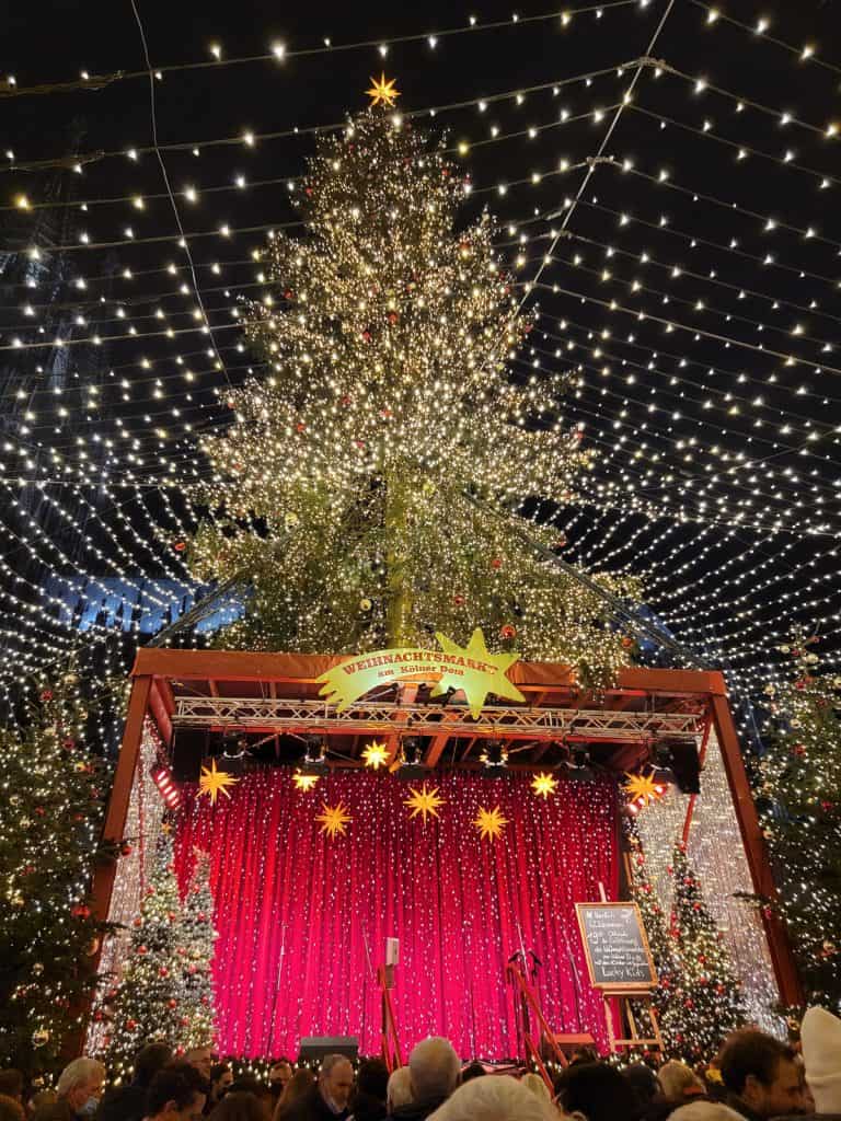 A very large wooden stage with a red background and 4 decorated Christmas trees stand in front of a huge Christmas trees with hundreds of lights.