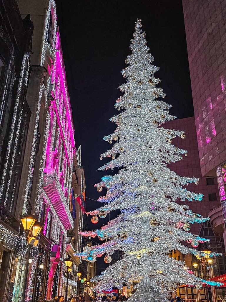 A large white lit up Christmas tree with the buildings around lit up with pink lights.