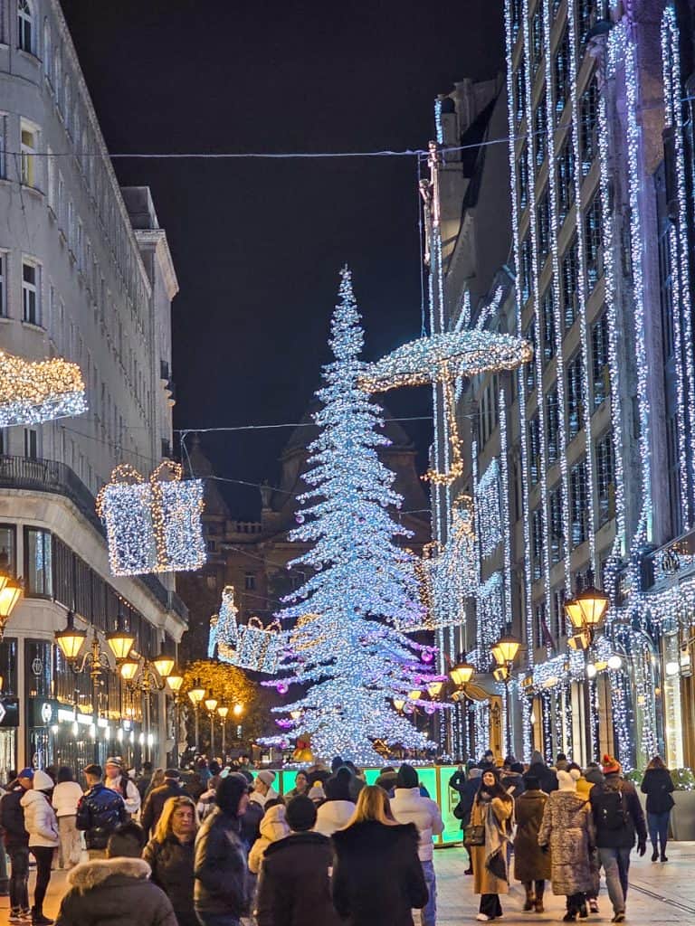 A lit up part of the market with a Christmas tree with blue lights and lit up presents being hung from side to side of the buildings