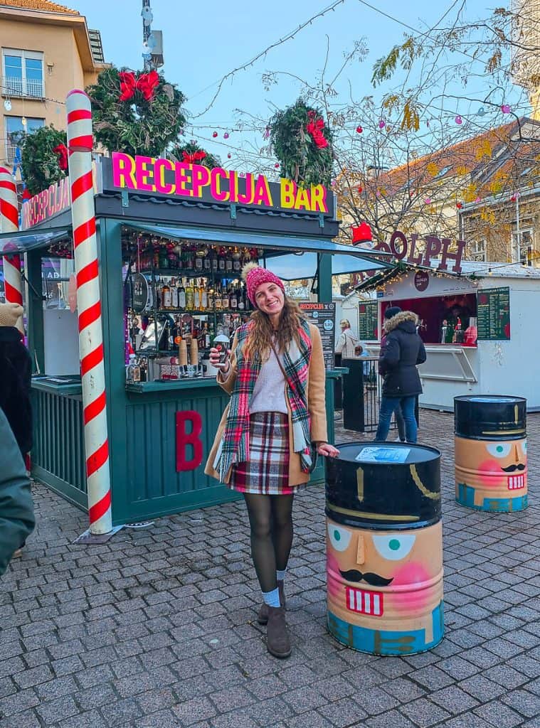 A woman holds a glass of hot cocoa in front of the wooden drink booth that is decorated with wreaths. There are lots of little tables around to hold your drinks that are painted to look like nutcrackers.