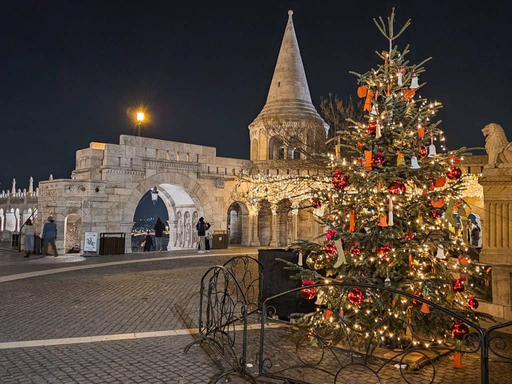A Christmas tree decorated with candles and small clay bells sits in front of a stone spire and wall.