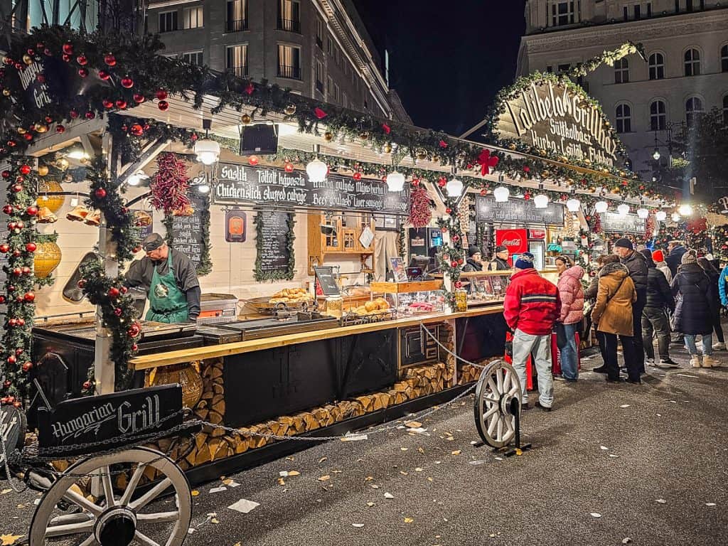 A drink and food station with wood stacked to the side and garlands. There are lots of food like bread, potatoes, and food with people lining up for a while.