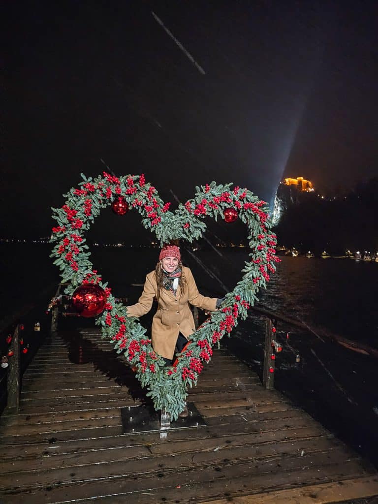 A large 10ft wreath shaped like a heart with red berries decorating it. A woman is standing inside the wreath that is located on the dock in front of the lake.