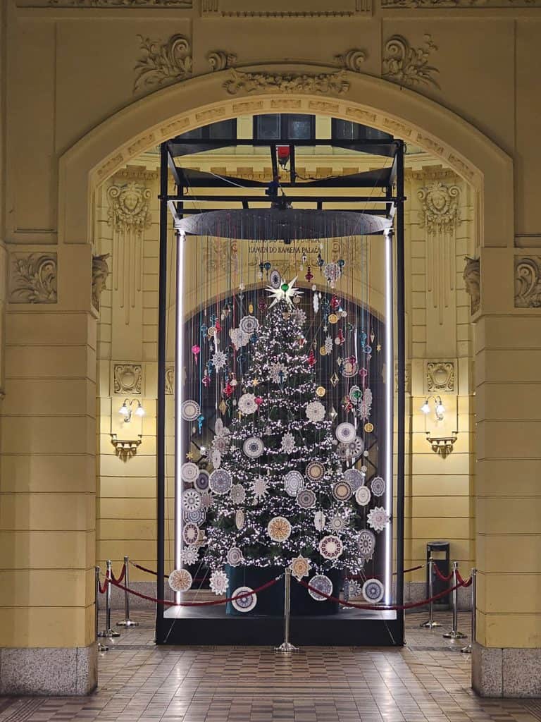 A Christmas tree set up in the middle of the cobblestone sidewalk is decorated by strands of paper decoratoin.