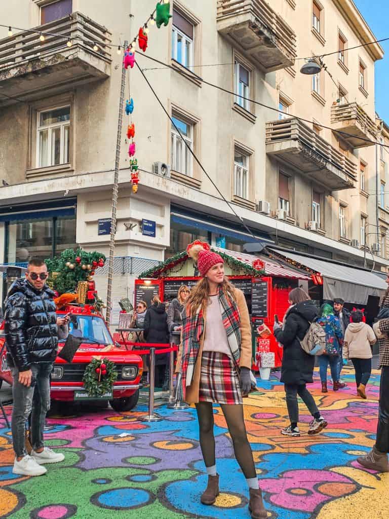 A woman stands in the middle of the street that is covered in a colorful cat mural.Behind the woman there is a red wooden drink stand and a red mustang decorated with a wreath.