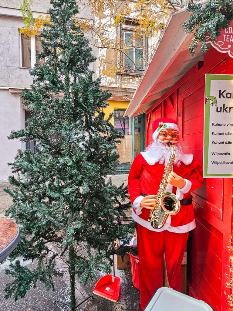 A Santa Claus holds a saxophone close to a scrawny green Christmas tree next to a red building.