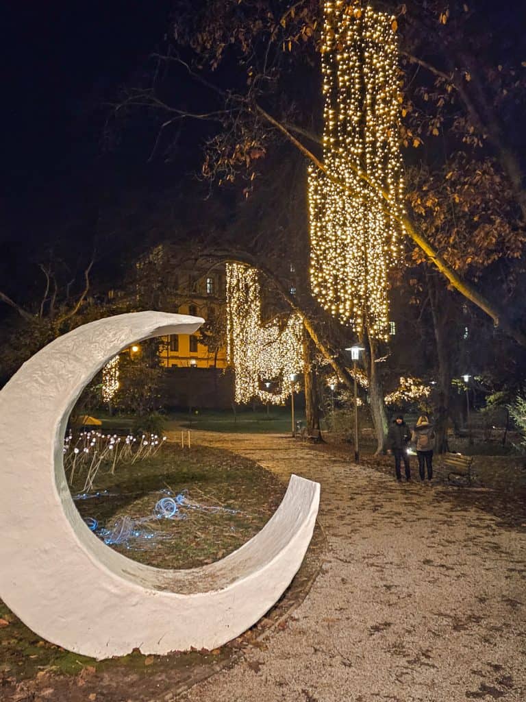 A large stone cresent moon on the side of the sidewalk in a park with lots of strings of light.