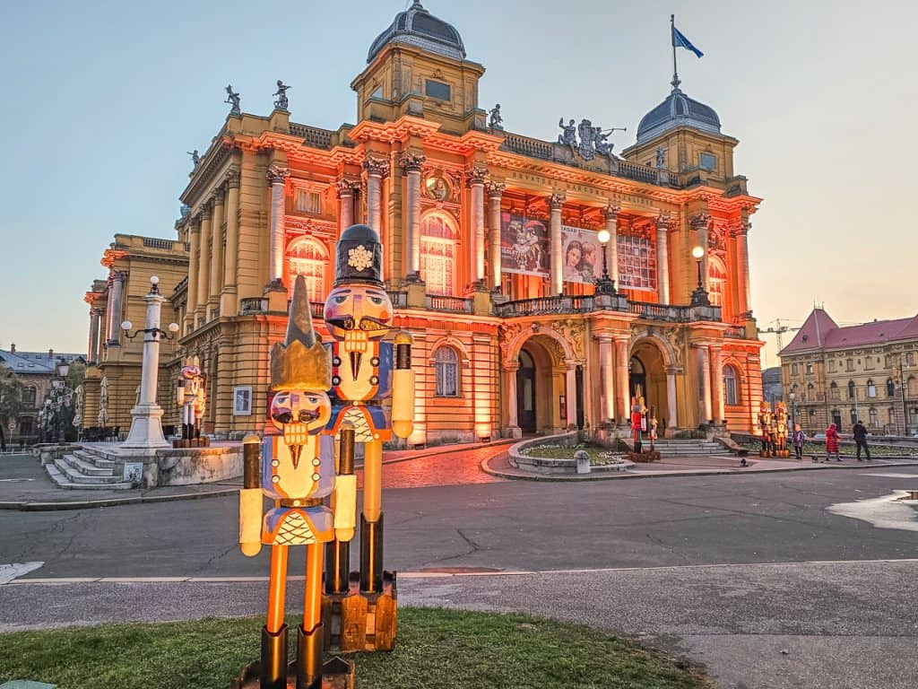 2 lit up nutcrackers stand outside the Zagreb National Theater that is lit up with orange lights.