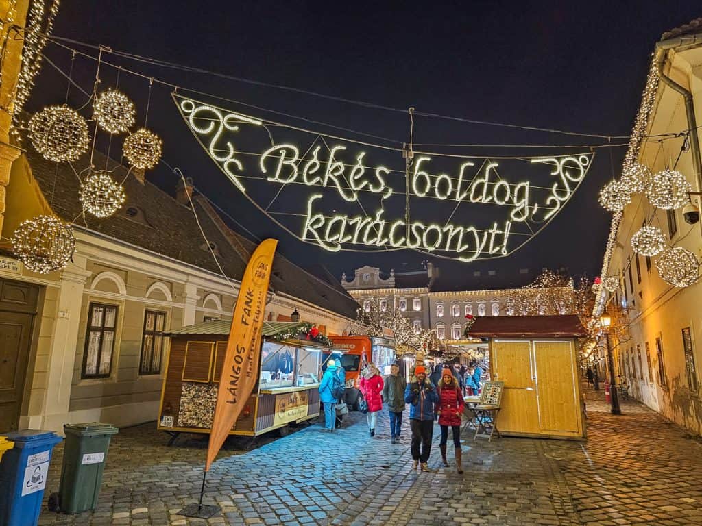 An alley with lots of wooden booths and lights hanging from the buildings saying Merry Christmas in Hungarian