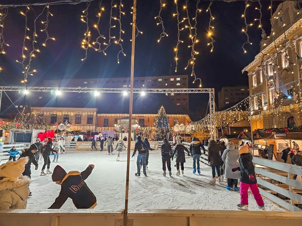 A ice rink with lots of people skating and a wooden fence around the edge with icicle lights hung around the edge.