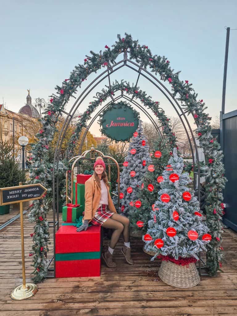A woman sits on a present with 3 Christmas trees next to her with an arched gateway (decorated with garlands) sits over head.