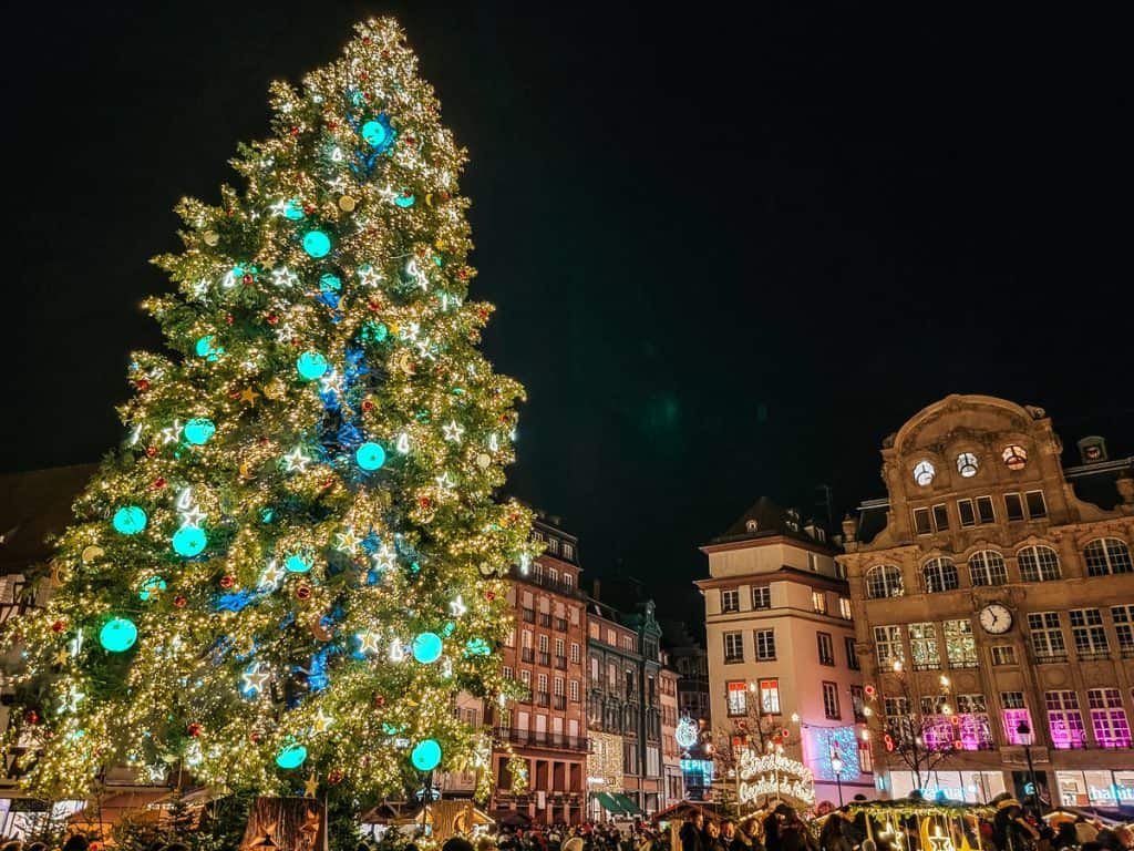A lit up christmas tree with lots of neon blue ornaments stands in the middle of the market square with an old stone clock tower to the side.