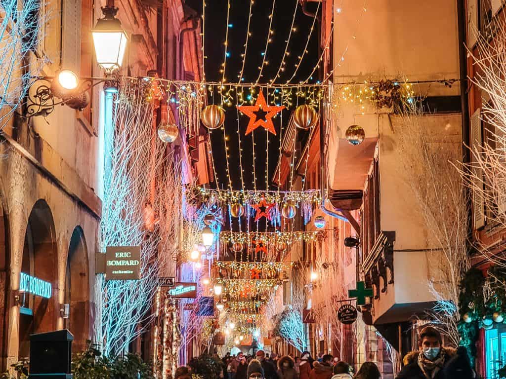 An alley with lights strung all the way down and paper stars being hung from the lights. Lots of people are walking around the yellow / tan-ish buildings.