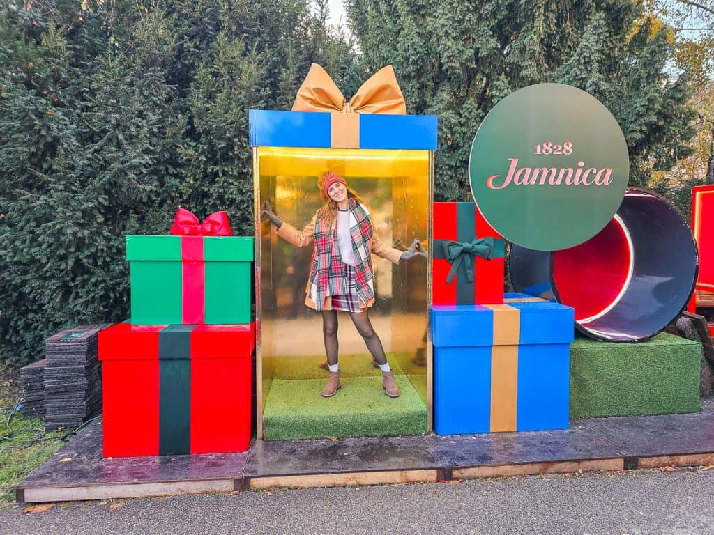 A woman stands inside of a blue wooden present with the front cut open. There are other presents around stacked up in different colors.