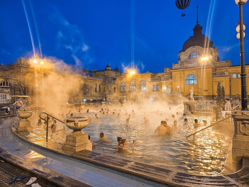 A large pool with lots of people swimming in it and fog covering some of the edges sits in front of a gorgeous arched building.