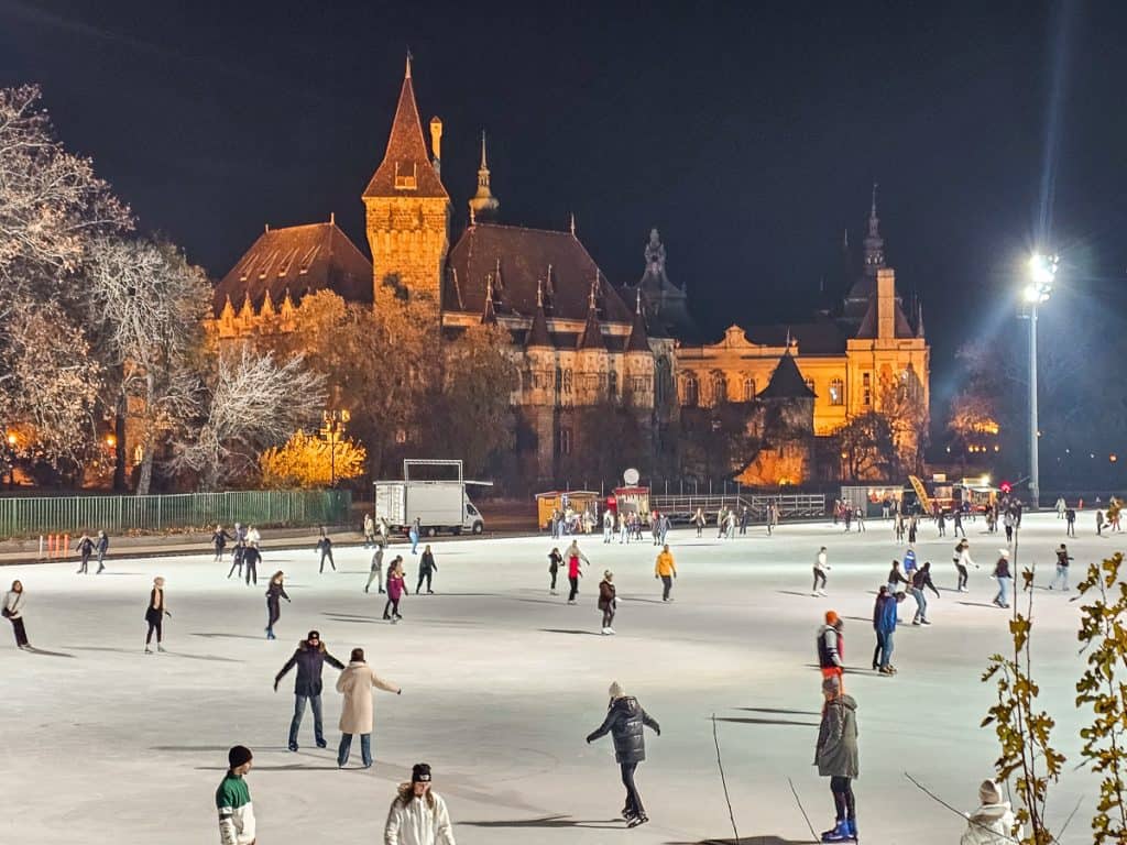 An ice skating rink with lots of people on it with a stone castle in the background. This is one of the best things to do in Budapest