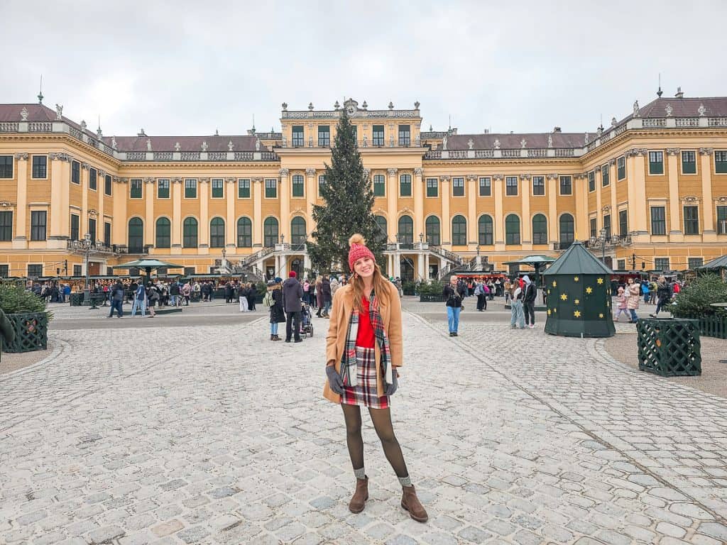 A woman in a tan coat and red sweater stands in front of the yellow and green elegant palace with Christmas trees and bushes lined all around the exterior.