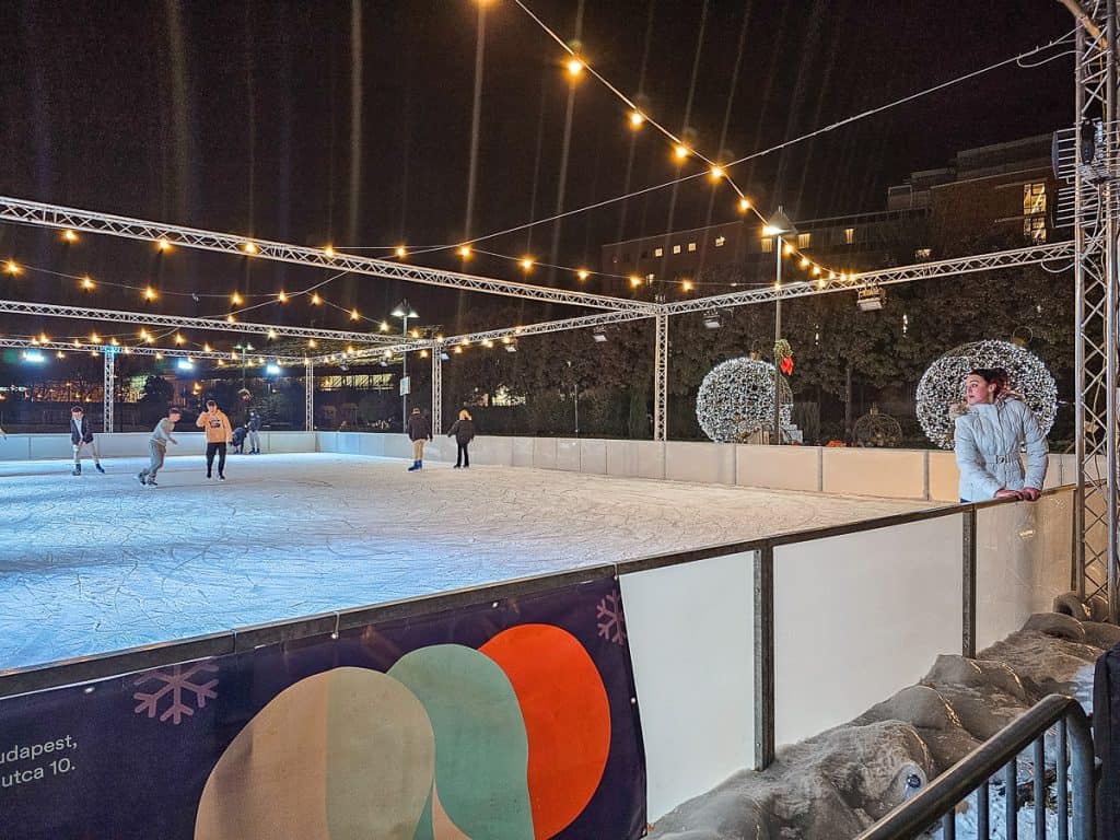 An ice skating rink with string lights going from side to side and a few people skating on the rink.