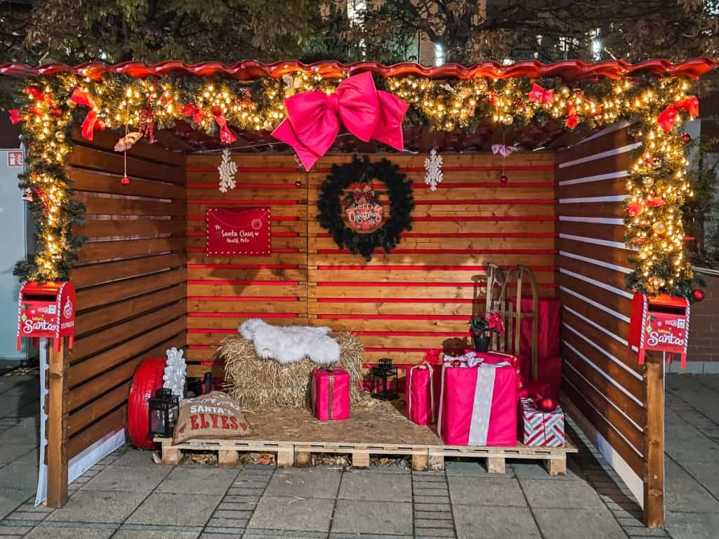 A small booth that is decorated with lights has a spot for pictures with red and white presents and a block of hay with a white blanket for people to sit on.