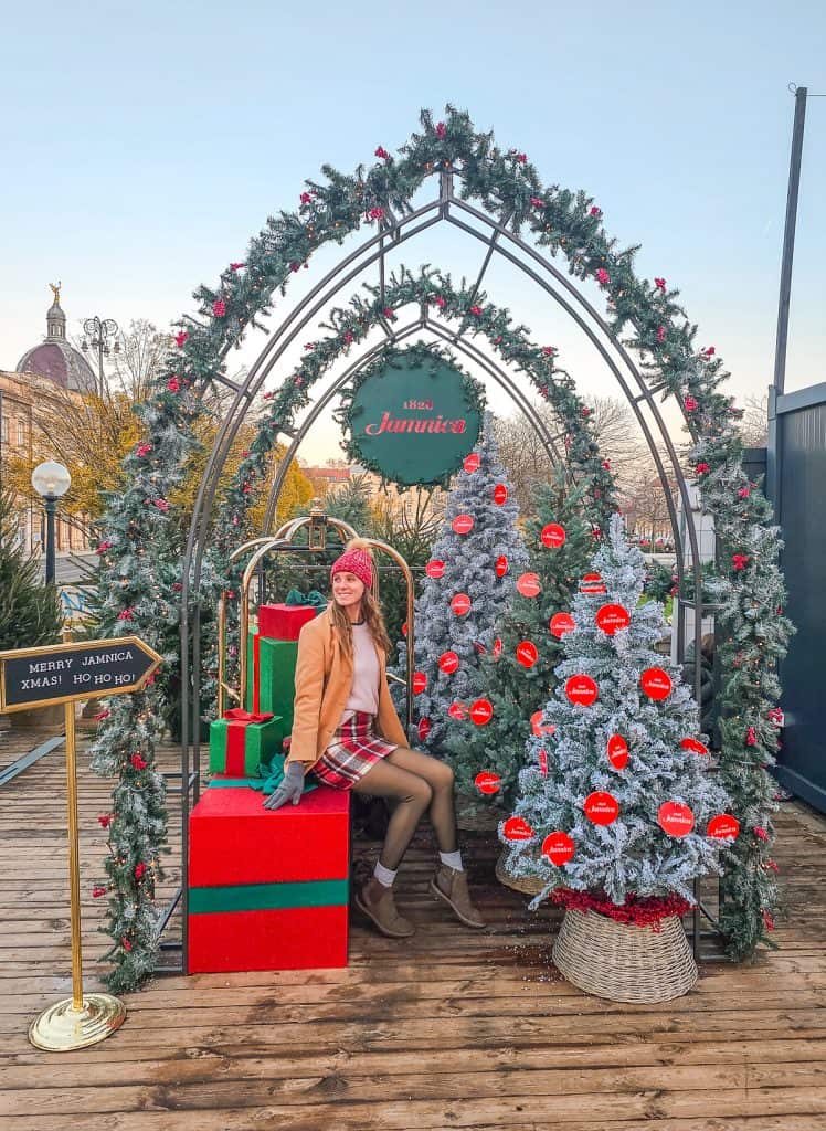A woman sits on a large red present with white and green christmas trees under an arched garland covered arched doorway.