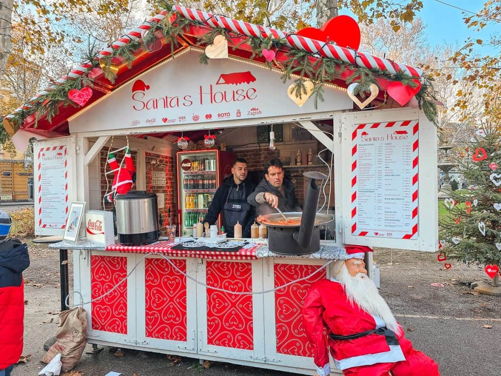 A small red booth (that has candy cane decoration and a Santa Claus that is standing outside ) that is serving assorted stews and hot chocolate.