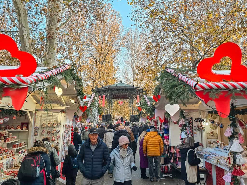 A gorgeous alley of the market with different stalls with red and white striped walls and decorated with garlands. There are lots of people walking around and heading up to the pavilion at the front of the alley.